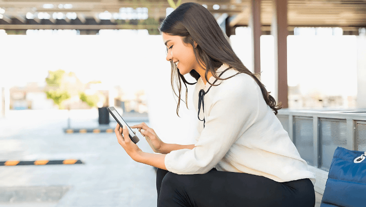 The Bright Future of E-Readers-This image depicts a young woman sitting outdoors in a modern setting, possibly at a bus stop or public seating area, using a tablet or e-reader.